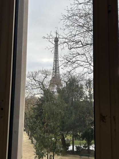 View of the Eiffel Tower framed by a window, with trees and a pathway visible in the foreground. The sky is overcast.